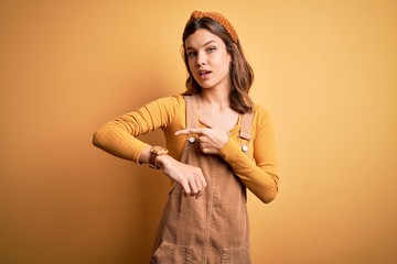 Young beautiful blonde girl wearing overall standing over yellow isolated background In hurry...