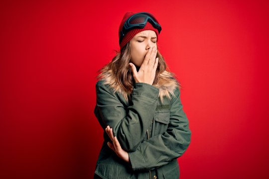 Young blonde girl wearing ski glasses and winter coat for ski weather over red background bored yawning tired covering mouth with hand. Restless and sleepiness.