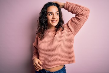 Young beautiful woman with curly hair wearing casual sweater over isolated pink background smiling confident touching hair with hand up gesture, posing attractive and fashionable