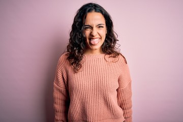 Fototapeta premium Young beautiful woman with curly hair wearing casual sweater over isolated pink background sticking tongue out happy with funny expression. Emotion concept.