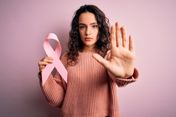 Beautiful woman with curly hair holding pink cancer ribbon symbol over isolated background with...