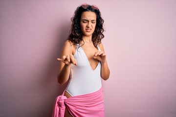 Beautiful woman with curly hair on vacation wearing white swimsuit over pink background disgusted...