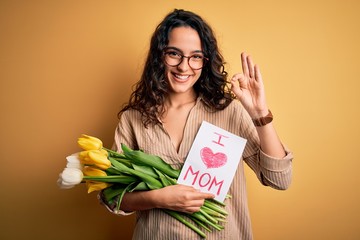 Beautiful curly hair woman holding love mom message and tulips celebrating mothers day doing ok sign with fingers, excellent symbol