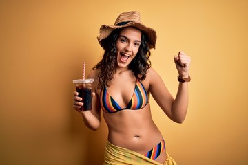 Young beautiful woman with curly hair on vacation wearing bikini drinking cola beverage screaming proud and celebrating victory and success very excited, cheering emotion