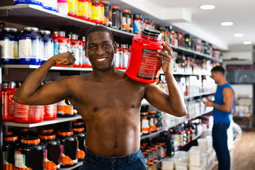 Fototapeta na wymiar Bare-chested athletic African standing with cans of sports nutrition in store, demonstrating muscles