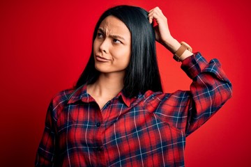 Young beautiful chinese woman wearing casual shirt over isolated red background confuse and wondering about question. Uncertain with doubt, thinking with hand on head. Pensive concept.