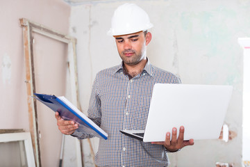 Professional constructor is standing with laptop and folder with documents