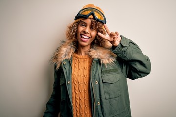 Young african american skier woman with curly hair wearing snow sportswear and ski goggles Doing peace symbol with fingers over face, smiling cheerful showing victory
