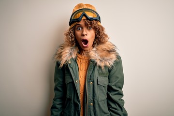 Young african american skier woman with curly hair wearing snow sportswear and ski goggles afraid and shocked with surprise and amazed expression, fear and excited face.