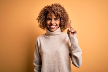 Young beautiful african american woman wearing turtleneck sweater over yellow background showing and pointing up with finger number one while smiling confident and happy.