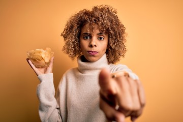 Naklejka premium Young african american curly woman holding bowl with chips potatoes over yellow background pointing with finger to the camera and to you, hand sign, positive and confident gesture from the front