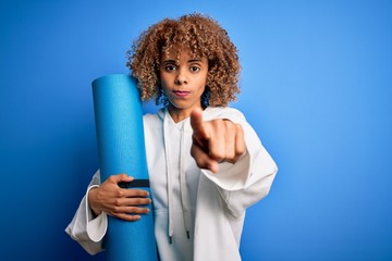 Young african american woman doing sport holding yoga mat over isolated blue background pointing with finger to the camera and to you, hand sign, positive and confident gesture from the front