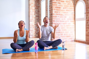 Fototapeta premium Middle age sporty couple sitting on mat doing stretching yoga exercise at gym crazy and mad shouting and yelling with aggressive expression and arms raised. Frustration concept.