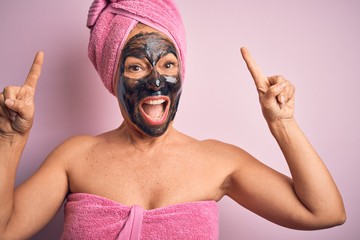 Middle age brunette woman wearing beauty black face mask over isolated pink background smiling amazed and surprised and pointing up with fingers and raised arms.