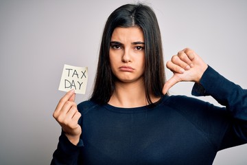 Young beautiful brunette woman holding paper with reminder paper with tax day message with angry...