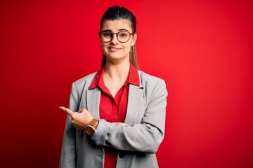 Young beautiful brunette businesswoman wearing jacket and glasses over red background Pointing aside worried and nervous with forefinger, concerned and surprised expression