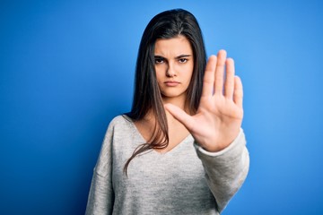 Young beautiful brunette woman wearing casual sweater standing over blue background doing stop sing...