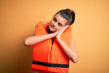 Young beautiful brunette woman wearing orange safe lifejacket over yellow background sleeping tired...