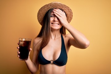 Young beautiful woman with blue eyes on vacation wearing bikini and hat drinking cola beverage stressed with hand on head, shocked with shame and surprise face, angry and frustrated. Fear and upset