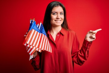 Young beautiful patriotic woman with blue eyes holding united states flags over red background very happy pointing with hand and finger to the side