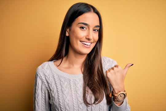 Young beautiful brunette woman wearing white casual sweater over yellow background smiling with happy face looking and pointing to the side with thumb up.