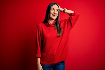 Young beautiful brunette woman wearing casual sweater over isolated red background smiling confident touching hair with hand up gesture, posing attractive and fashionable