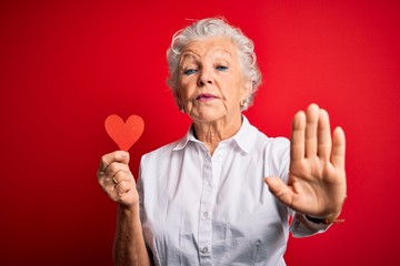 Senior beautiful woman holding paper heart standing over isolated red background with open hand doing stop sign with serious and confident expression, defense gesture