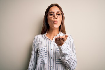 Young beautiful brunette woman wearing casual shirt and glasses over white background looking at the camera blowing a kiss with hand on air being lovely and sexy. Love expression.