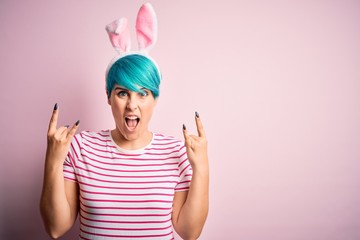 Obraz na płótnie Canvas Young woman with fashion blue hair wearing easter rabbit ears over pink background shouting with crazy expression doing rock symbol with hands up. Music star. Heavy concept.