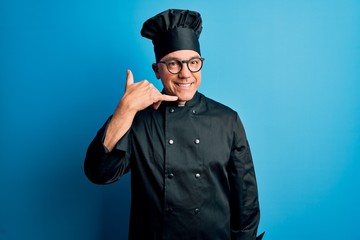 Middle age handsome grey-haired chef man wearing cooker uniform and hat smiling doing phone gesture with hand and fingers like talking on the telephone. Communicating concepts.