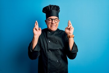 Middle age handsome grey-haired chef man wearing cooker uniform and hat gesturing finger crossed...