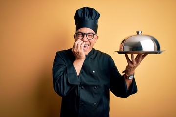 Middle age handsome grey-haired waiter man wearing cooker uniform and hat holding tray laughing and...