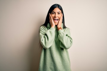 Young beautiful hispanic woman wearing green winter sweater over isolated background afraid and shocked, surprise and amazed expression with hands on face