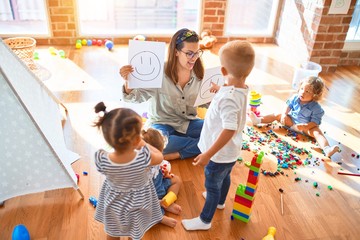 Beautiful psychologist and group of toddlers make therapy using emotions emojis around lots of toys at kindergarten - obrazy, fototapety, plakaty