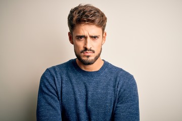 Young handsome man with beard wearing casual sweater standing over white background skeptic and...