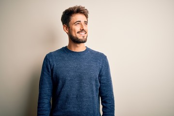 Young handsome man with beard wearing casual sweater standing over white background looking away to...