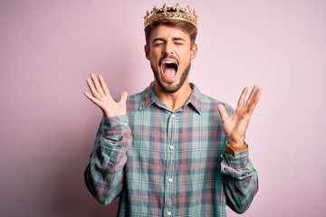 Young man with beard wearing golden crown of king standing over isolated pink background...