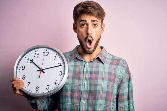 Young man with beard doing countdown using big clock over isolated pink background scared in shock with a surprise face, afraid and excited with fear expression
