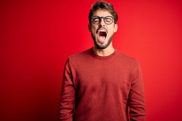 Fototapeta na wymiar Young handsome man with beard wearing glasses and sweater standing over red background angry and mad screaming frustrated and furious, shouting with anger. Rage and aggressive concept.