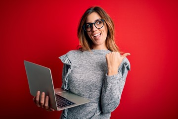 Naklejka premium Young beautiful brunette businesswoman wearing glasses using laptop over red background pointing and showing with thumb up to the side with happy face smiling