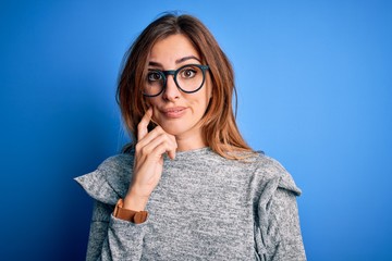 Young beautiful brunette woman wearing casual sweater and glasses over blue background with hand on chin thinking about question, pensive expression. Smiling with thoughtful face. Doubt concept.