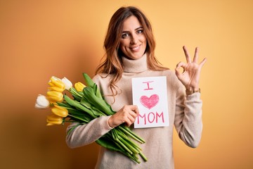 Beautiful brunette woman holding love mom message and tulips celebrating mothers day doing ok sign with fingers, excellent symbol