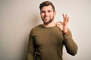 Young blond man with beard and blue eyes wearing green sweater over white background smiling positive doing ok sign with hand and fingers. Successful expression.
