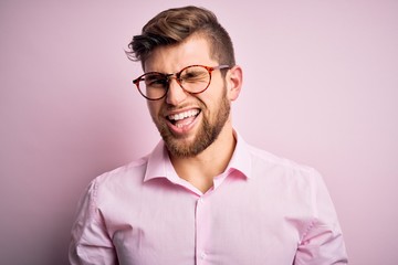 Young handsome blond man with beard and blue eyes wearing pink shirt and glasses winking looking at...
