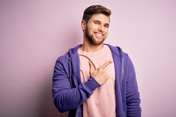 Young blond man with beard and blue eyes wearing purple sweatshirt over pink background cheerful with a smile of face pointing with hand and finger up to the side with happy and natural expression