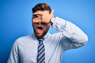 Young blond businessman with beard and blue eyes wearing elegant shirt and tie standing peeking in shock covering face and eyes with hand, looking through fingers with embarrassed expression.