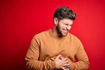 Naklejka premium Young blond man with beard and blue eyes wearing casual s over red background with hand on stomach because nausea, painful disease feeling unwell. Ache concept.