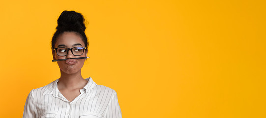 Black girl in glasses keeping pencil like mustache and looking aside
