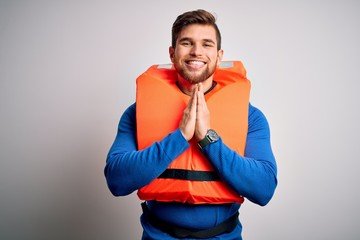 Young blond tourist man with beard and blue eyes wearing lifejacket over white background praying...