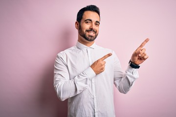 Young handsome man with beard wearing casual shirt standing over pink background smiling and looking at the camera pointing with two hands and fingers to the side.